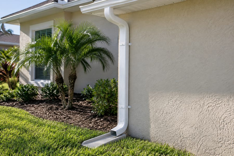 White aluminum downspout strapped to a stucco wall, discharging onto a splash block by a palm bed